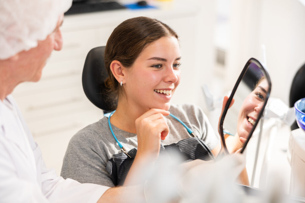 During appointment, dentist demonstrates to patient result of cosmetic dental filling. Senior man specialist holds mirror in front of girl patient and suggests evaluating effect of treatment.
