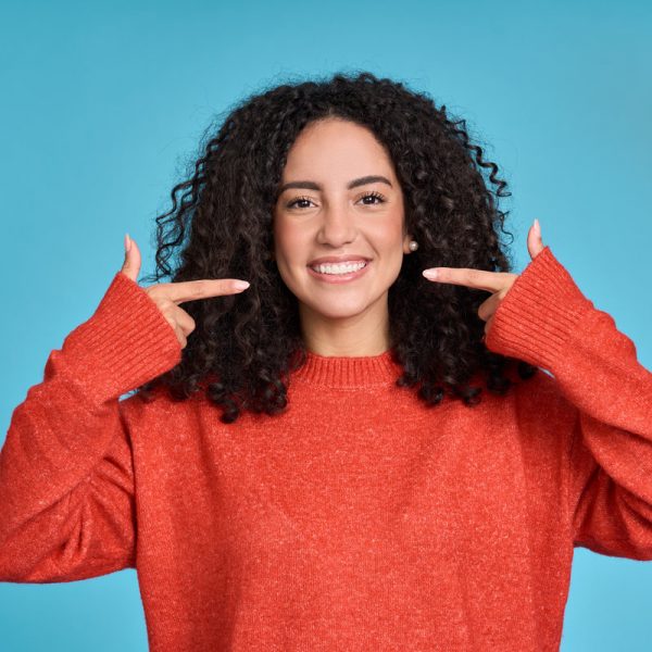 Happy young latin woman pointing at white healthy teeth, showing perfect dental toothy smile isolated on blue background advertising dentistry tooth care whitening clinic services concept.